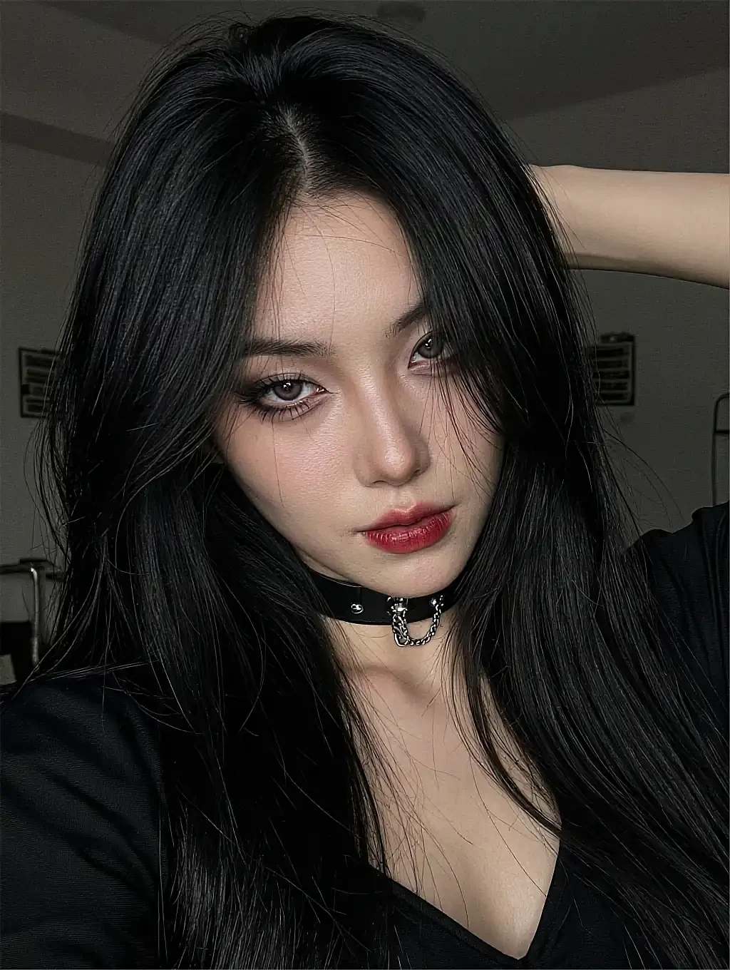 Dark-haired woman with red lips and a leather chain choker, close-up selfie in a dim room